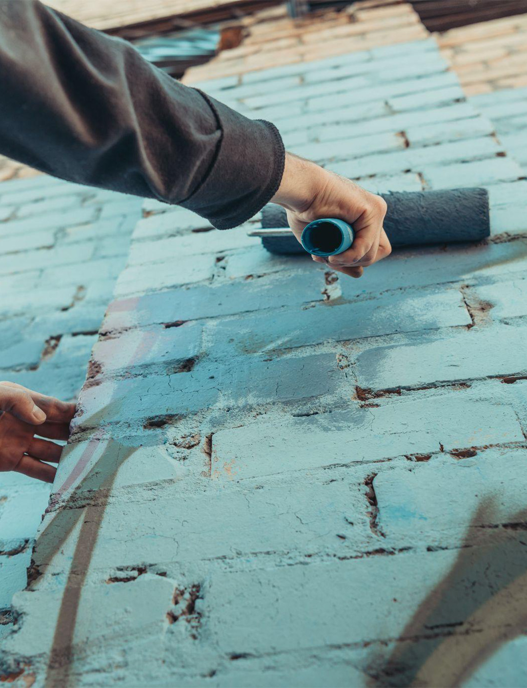 Graffiti-Removal-3 A person's hand using a small paint roller to apply light blue paint on a brick surface.